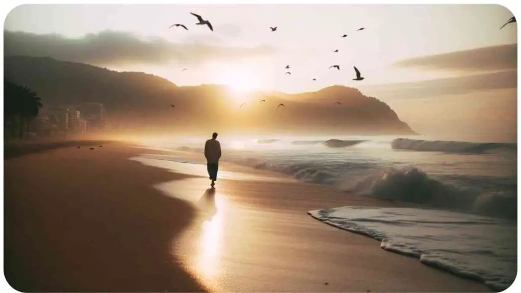 Photo of a peaceful beach during sunrise. A person of Hispanic descent is walking along the shore, feeling the wet sand beneath their feet and listening to the rhythmic sound of waves. Birds fly overhead, and the individual appears lost in the mindfulness of the moment.