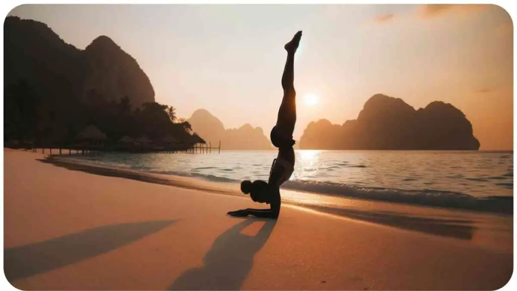 Photo of a serene beach setting during sunset. A person of African descent is gracefully executing the Downward Dog Pose (Adho Mukha Svanasana), forming an inverted V-shape with hands and feet firmly on the ground and hips raised.